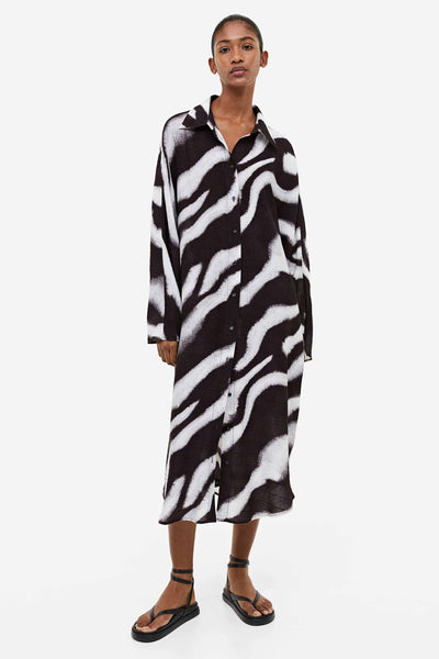 A model wearing a black and white zebra print long shirt dress with a collar, long sleeves, and flat black sandals.