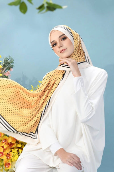Woman showcasing a sunshine yellow hijab with black polka dots and trim, complemented by a floral background for a fresh and fashionable look